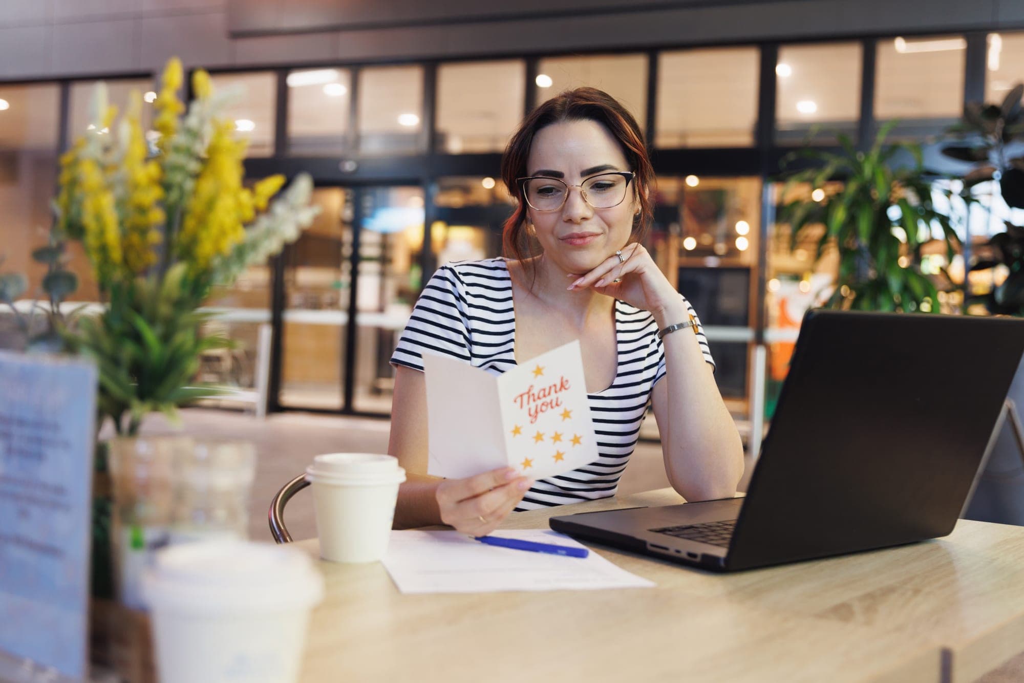 Woman at a café laptop smiling at a handwritten thank-you card