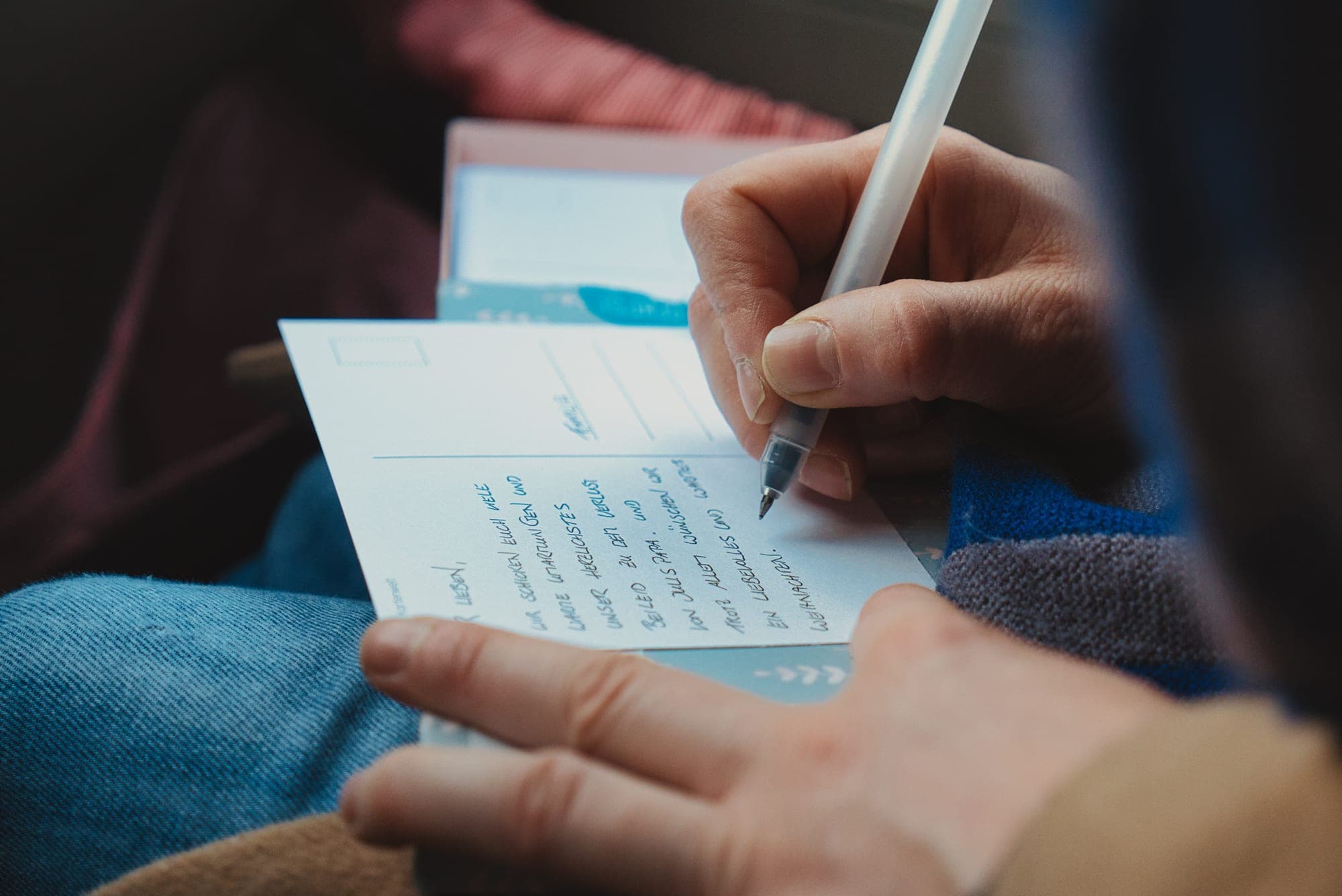A hand writing a message in blue ink on a postcard, the lines of handwriting filling the card