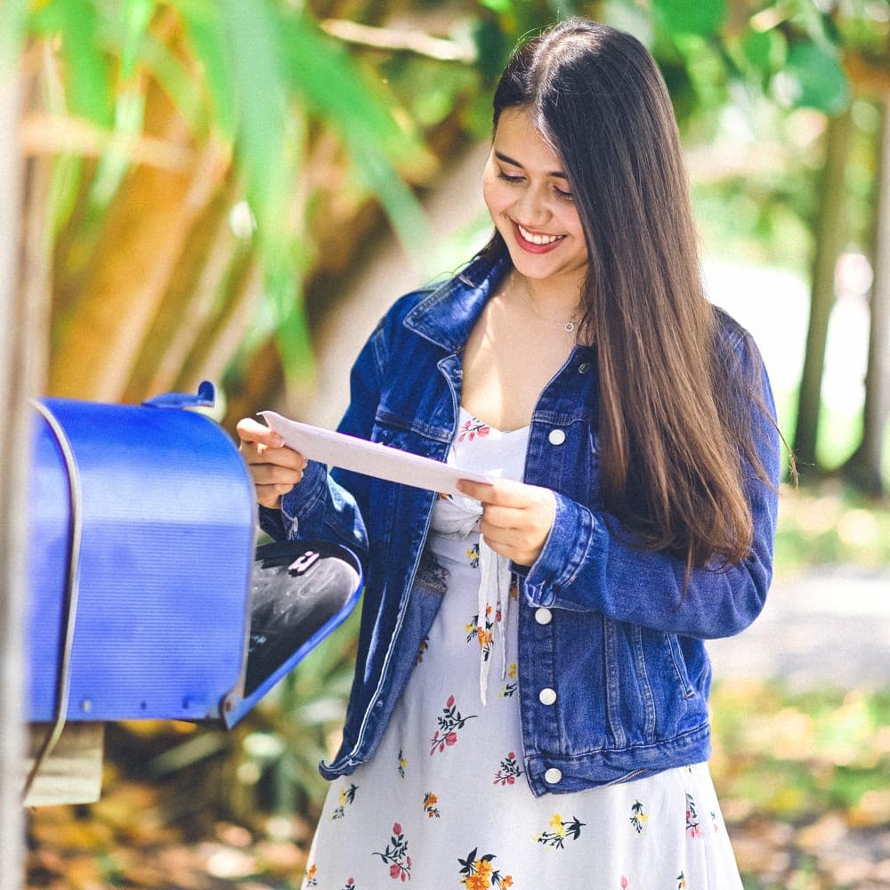 A smiling woman in a denim jacket reading a card she has just taken from a blue mailbox