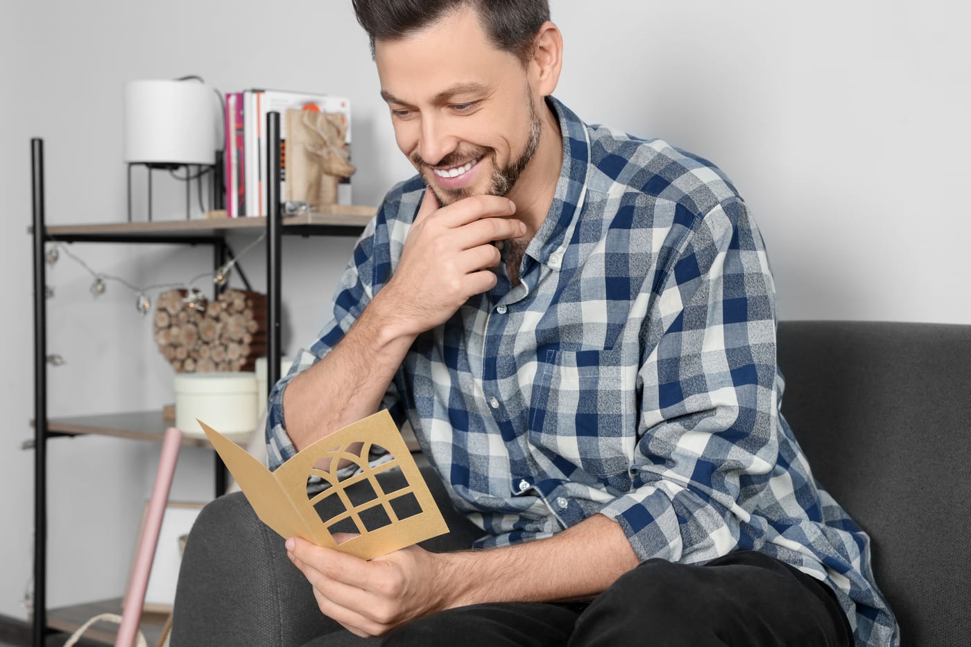 A smiling man in a checked shirt sitting on his sofa at home, reading a greeting card he has just opened