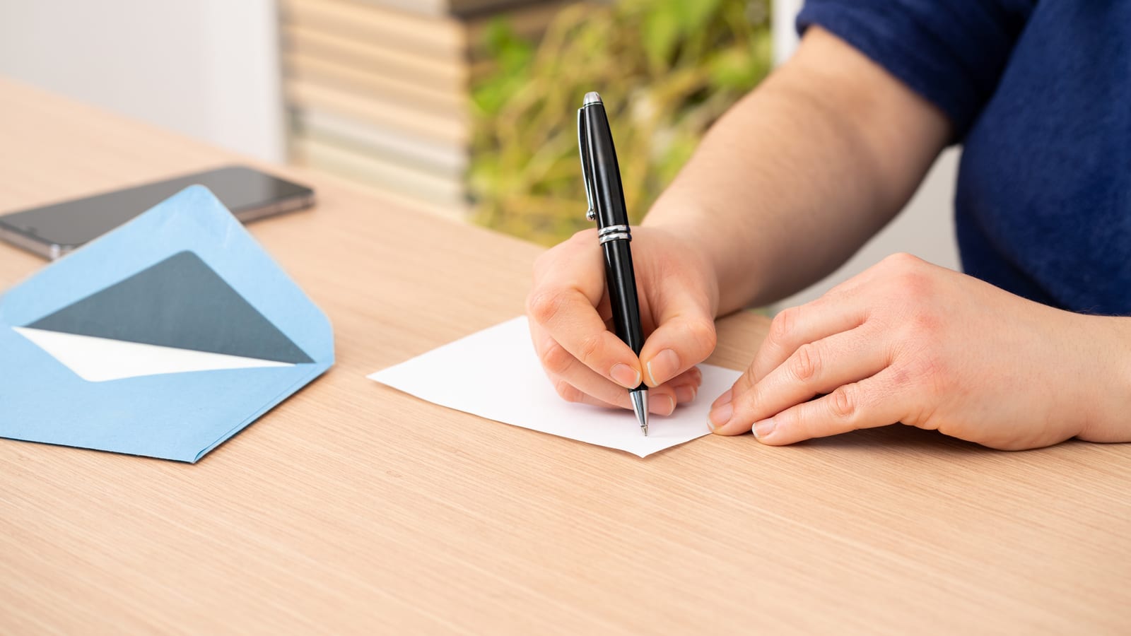 A woman at a wooden desk writing a thank-you card by hand with a black pen, a pale blue envelope beside it