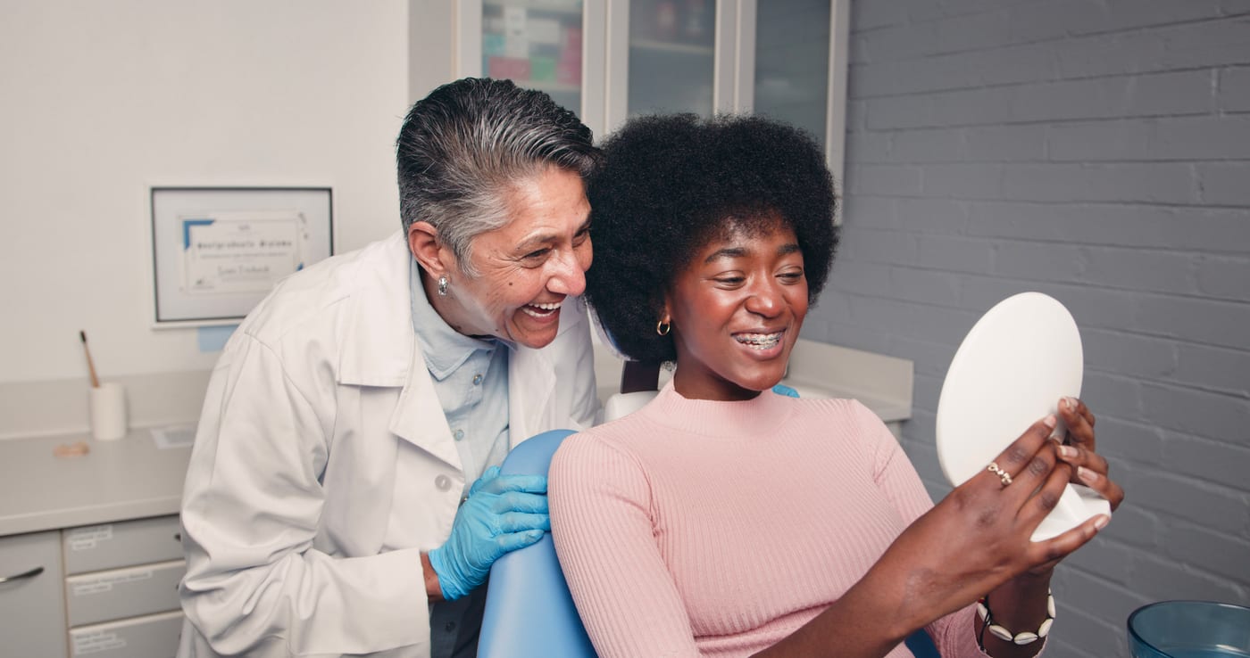 A young patient checking her new smile in a hand mirror while her orthodontist smiles beside her