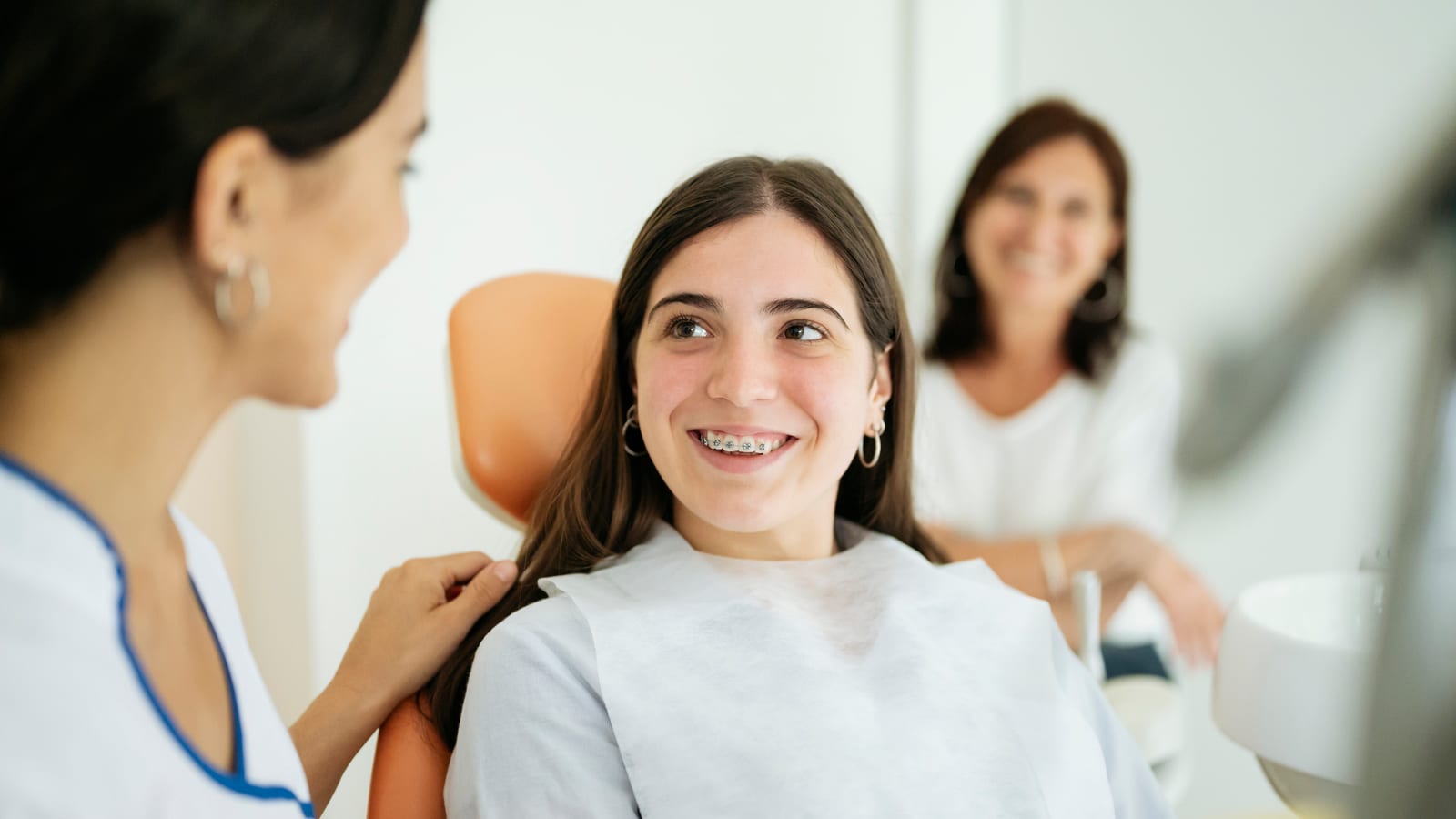 A smiling teenage girl in the orthodontist's chair talking with her orthodontist, her mother behind her