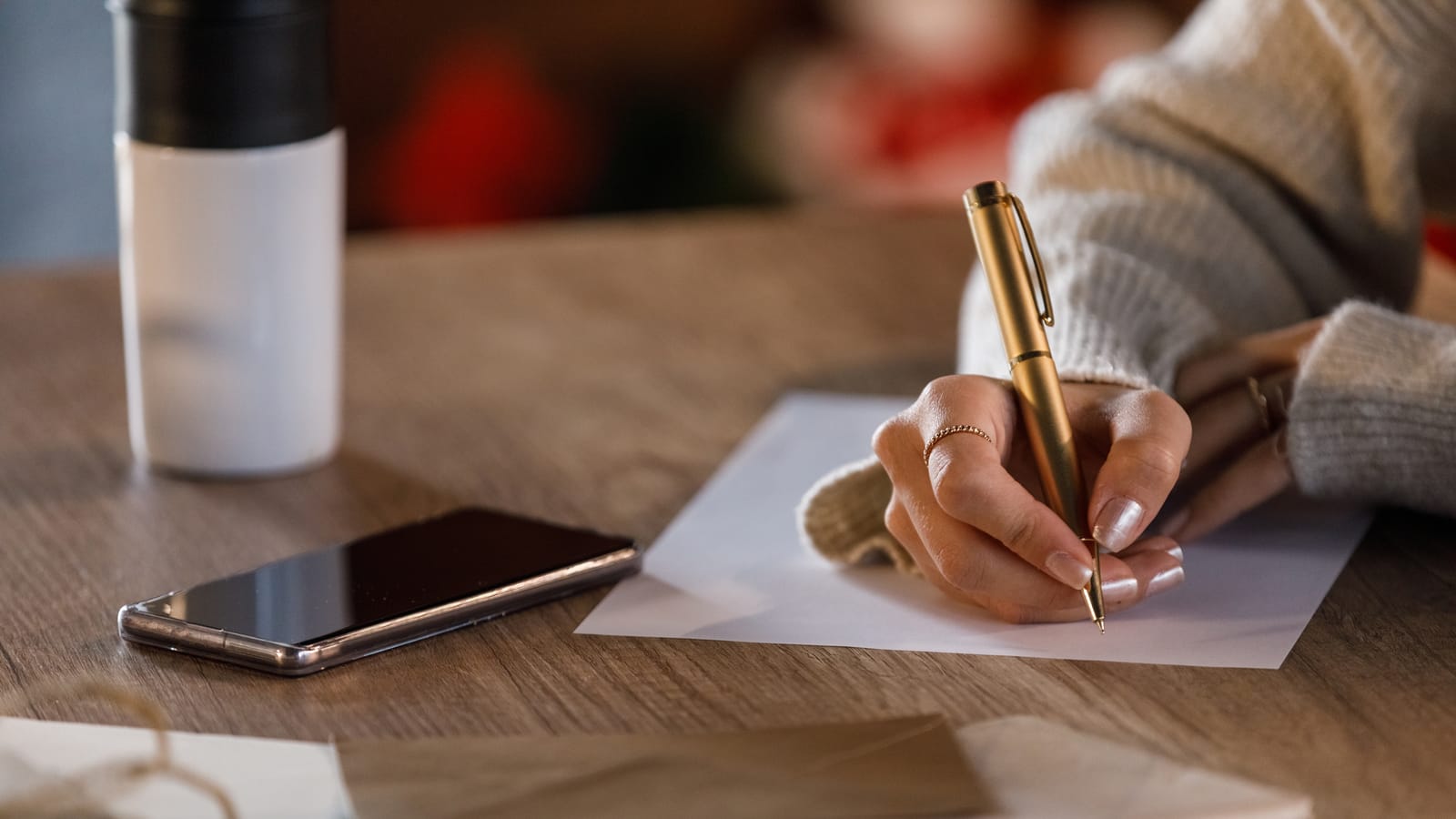 A woman in a cream sweater writing a holiday card with a gold pen at a wooden table, her phone and a cup beside her