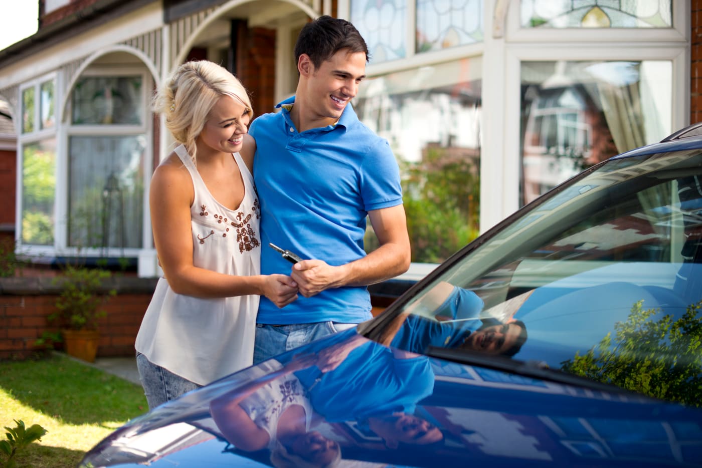 A young couple in their driveway reading a handwritten card together beside their new car, the dealer envelope in hand