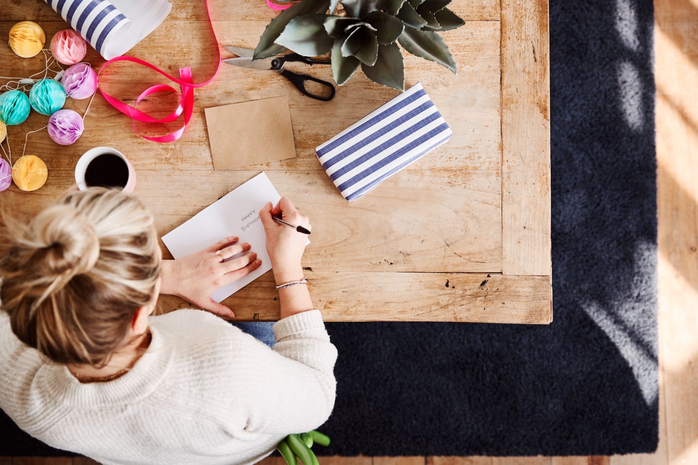 Looking down on a woman at a wooden table writing a birthday card, with ribbon, wrapping paper and a coffee beside her