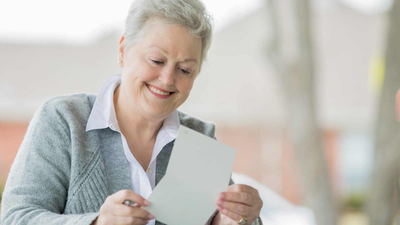 A smiling older woman with short grey hair reading a handwritten greeting card by a bright window at home