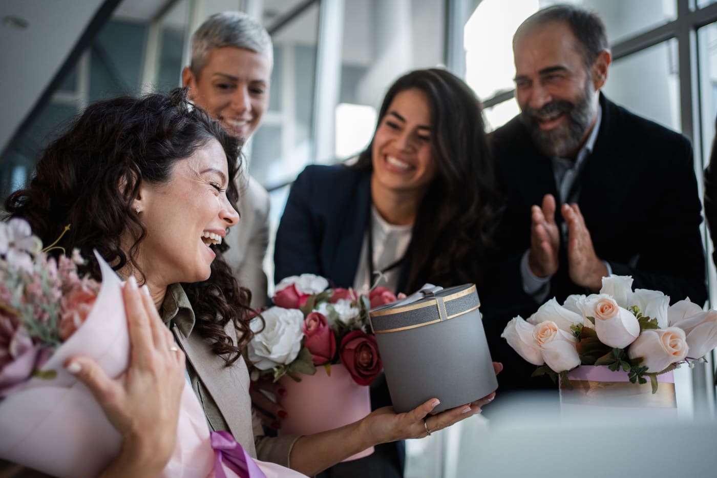 Colleagues clapping and laughing around a delighted coworker holding flowers and a gift box in a busy office
