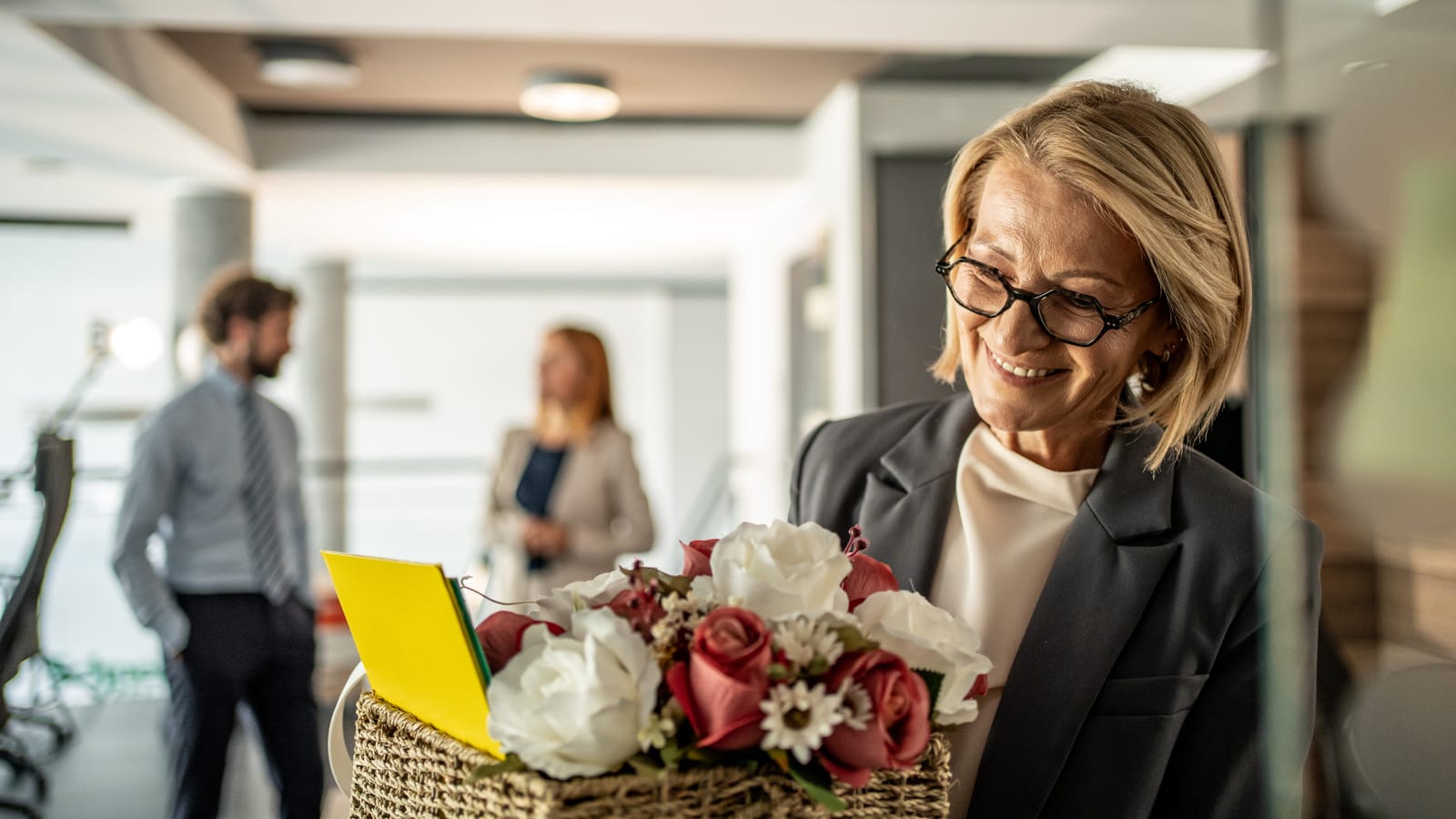 A smiling woman in glasses at the office opening a basket of flowers with a yellow card tucked in from her colleagues