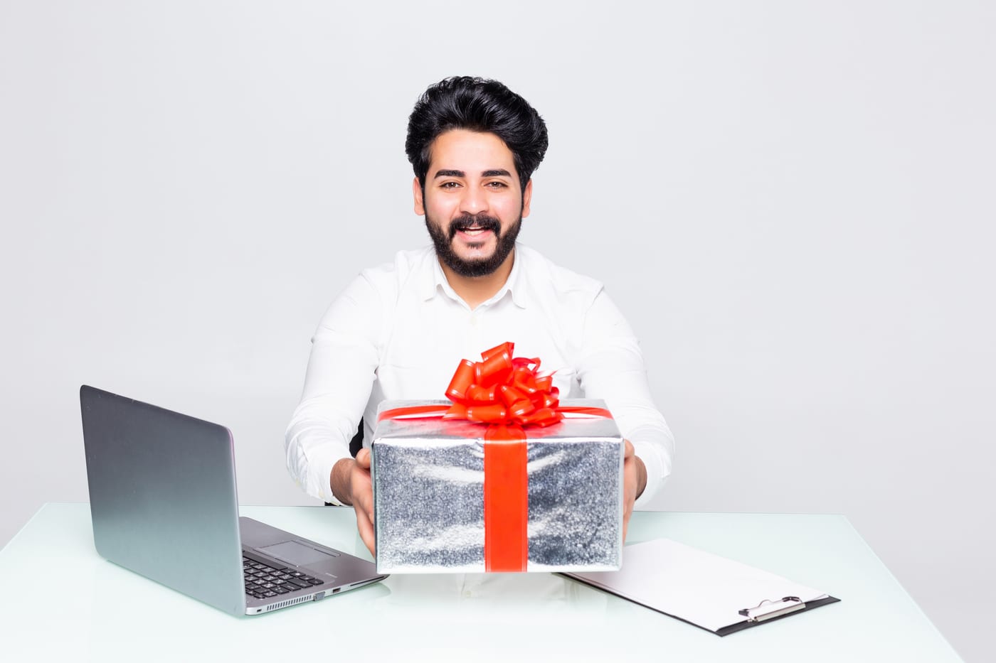 A smiling young businessman at his office desk holding a wrapped gift box with a red bow