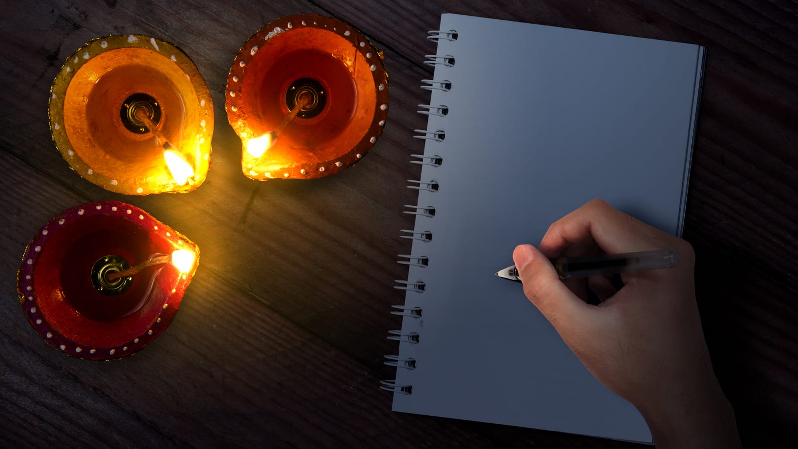 A hand writing in a spiral notebook beside three lit clay diyas glowing on a dark wooden table at Diwali