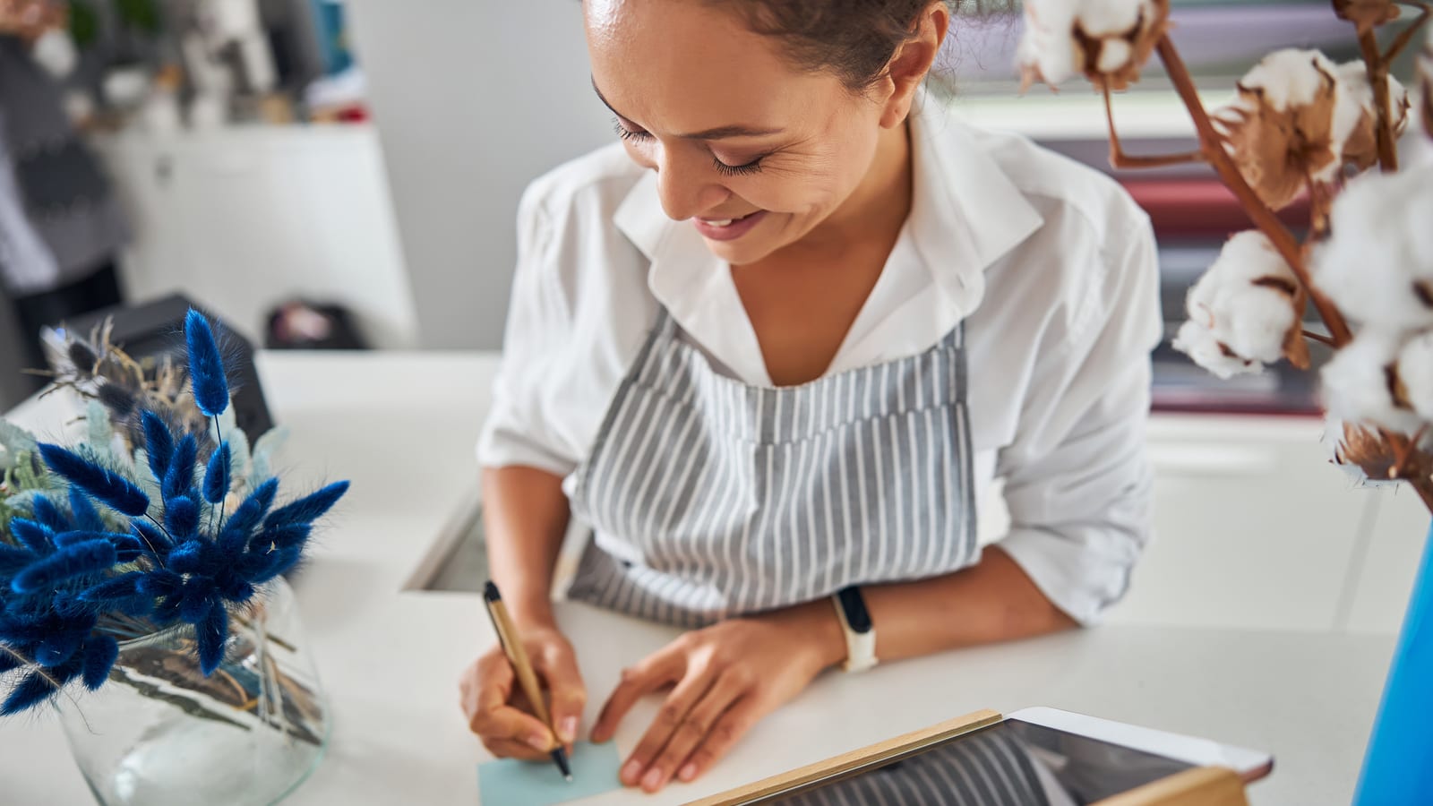 A shop owner in a striped apron at her counter, writing a card by hand with a pen