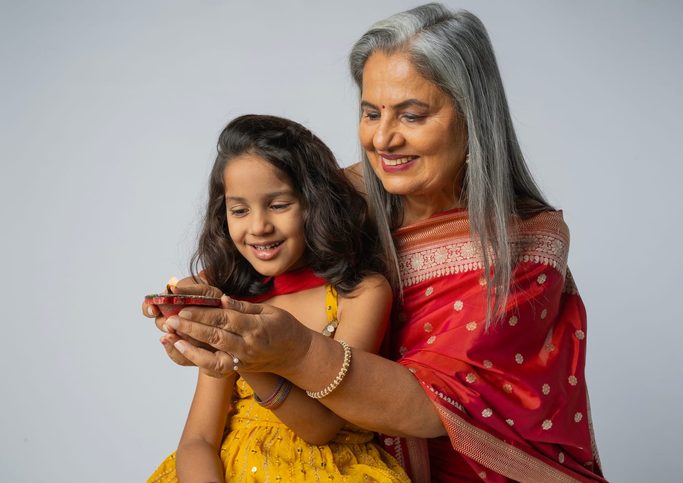 A grandmother in a red sari and her granddaughter holding a lit diya together, both smiling