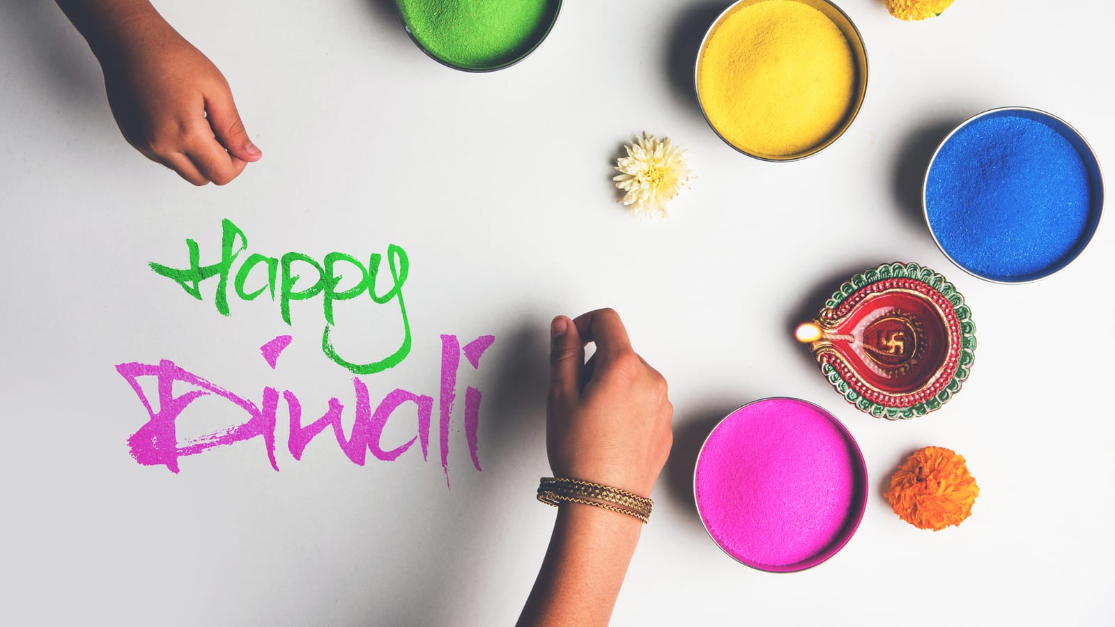 A girl writing Happy Diwali in coloured rangoli powder, bowls of bright colour and a lit diya around it