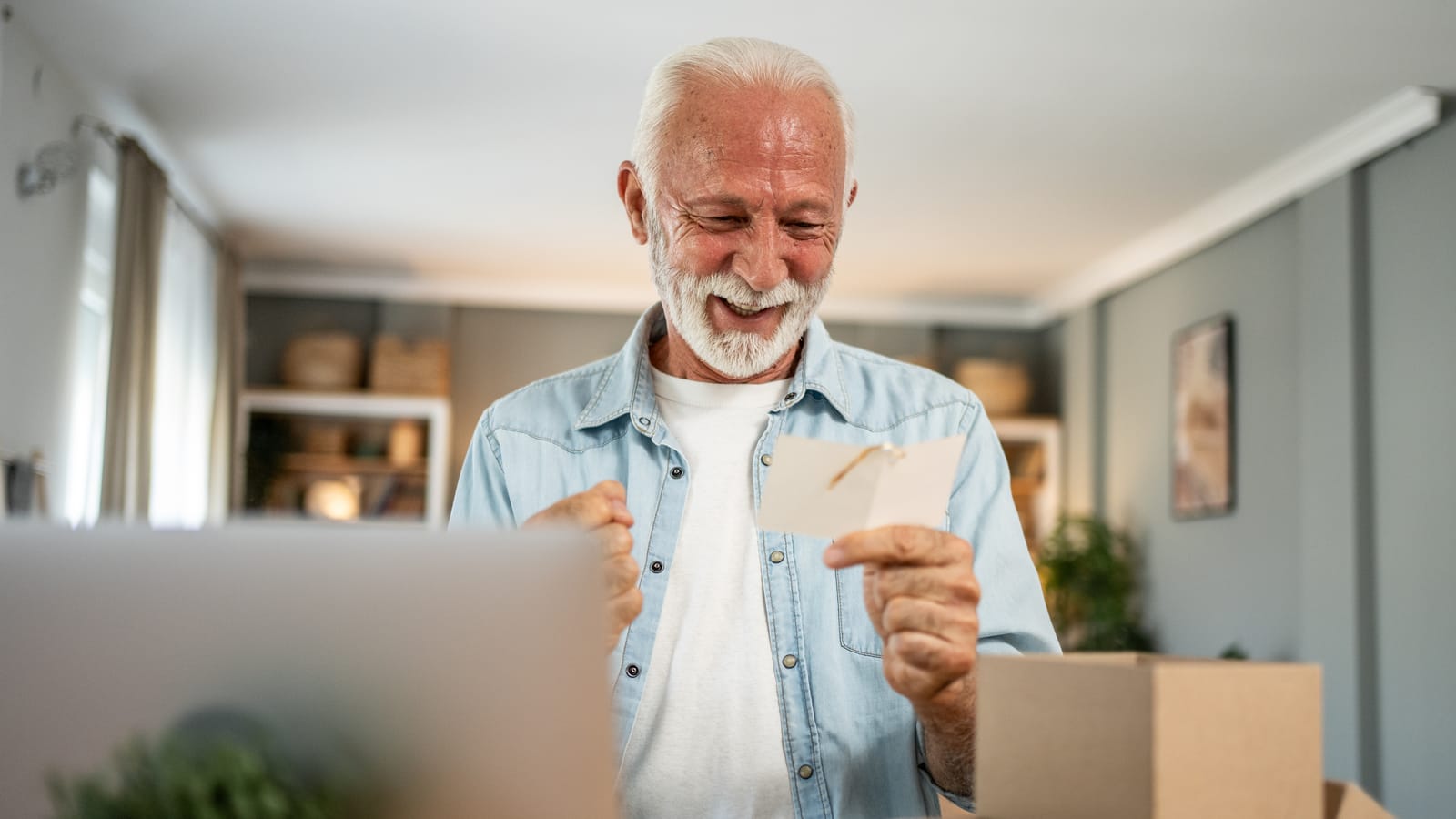 A smiling older man in a denim shirt reading a small card he has just taken out of a delivery box at home