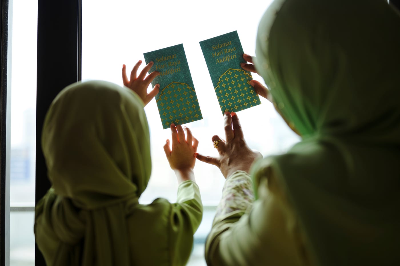 A mother and young daughter in green hijabs hanging green Eid money envelopes on a sunlit window