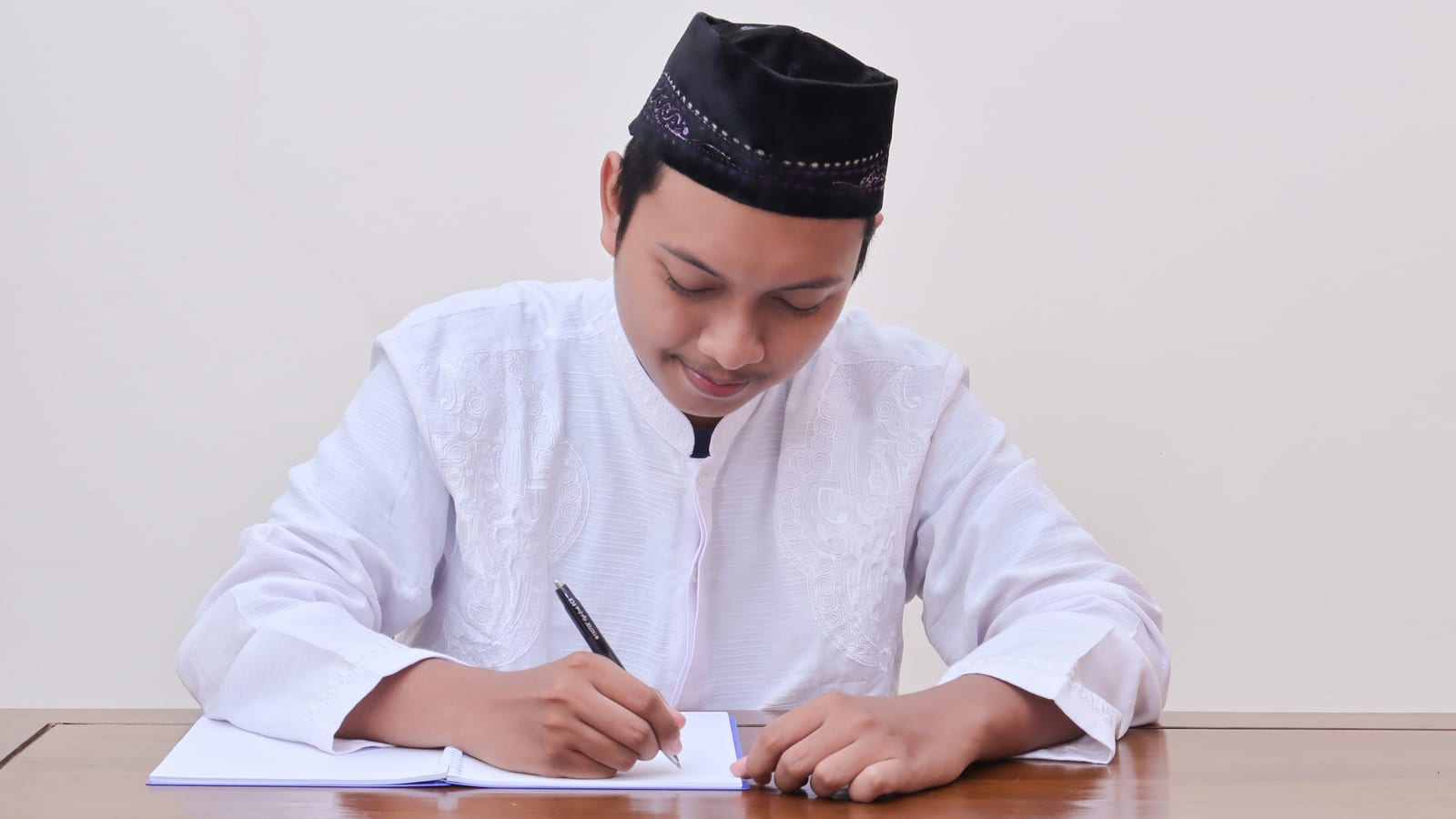 A young man in a white kurta and black cap sitting at a table, writing carefully by hand with a pen