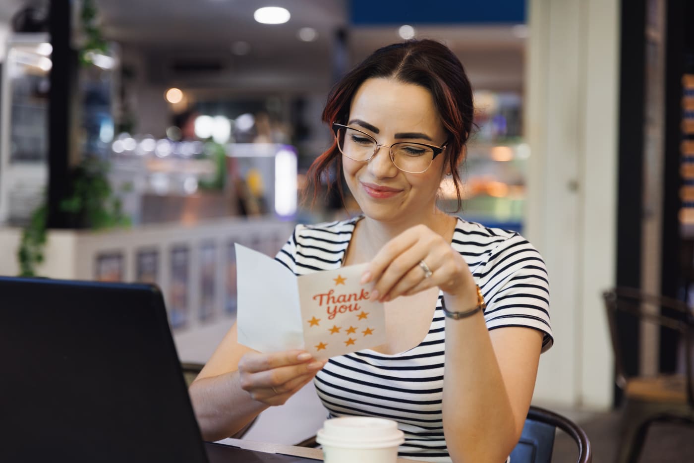A young woman in glasses smiling as she opens a Thank You card at her desk, her laptop open in front of her