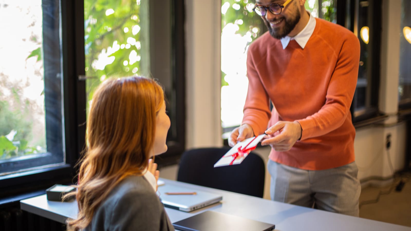 A man in an orange sweater handing a card and a small gift to a surprised colleague at her desk in a bright office