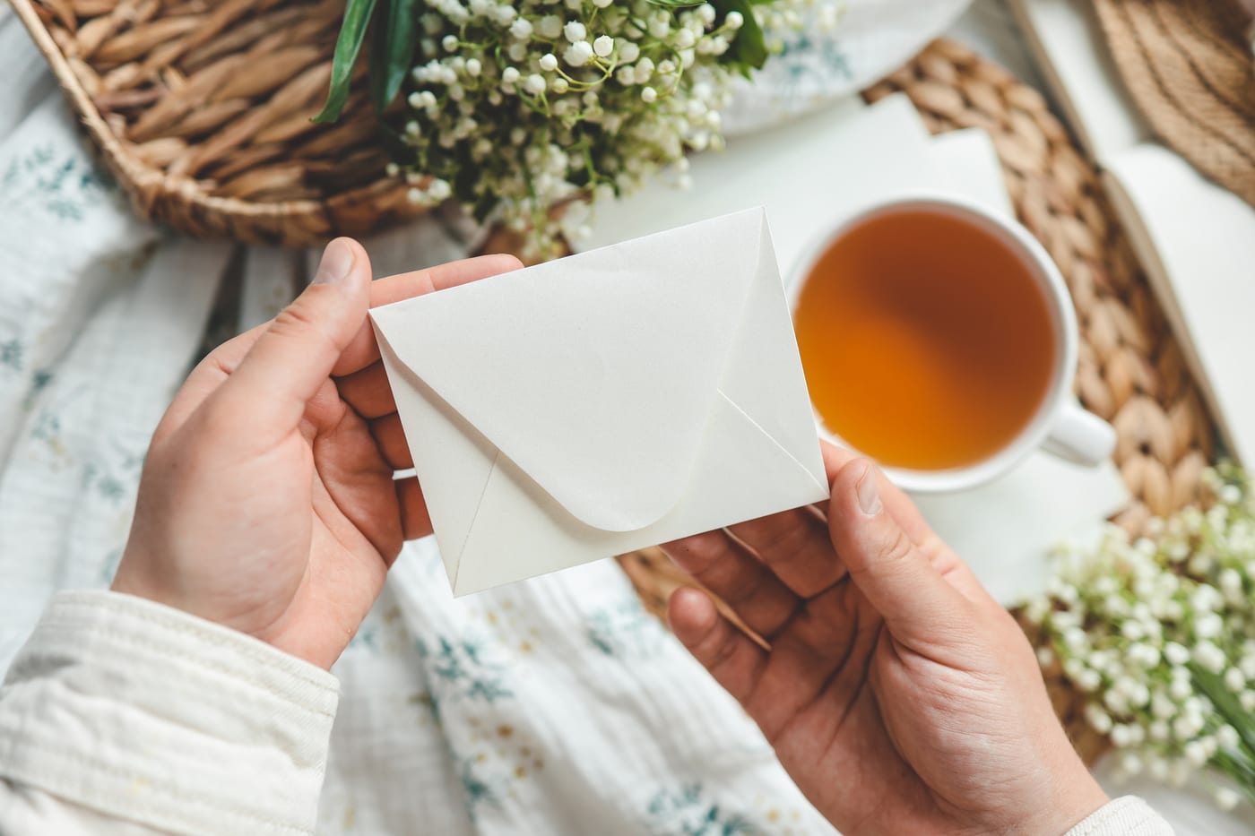Hands holding a cream envelope over a cup of tea, with small white flowers on the table around it