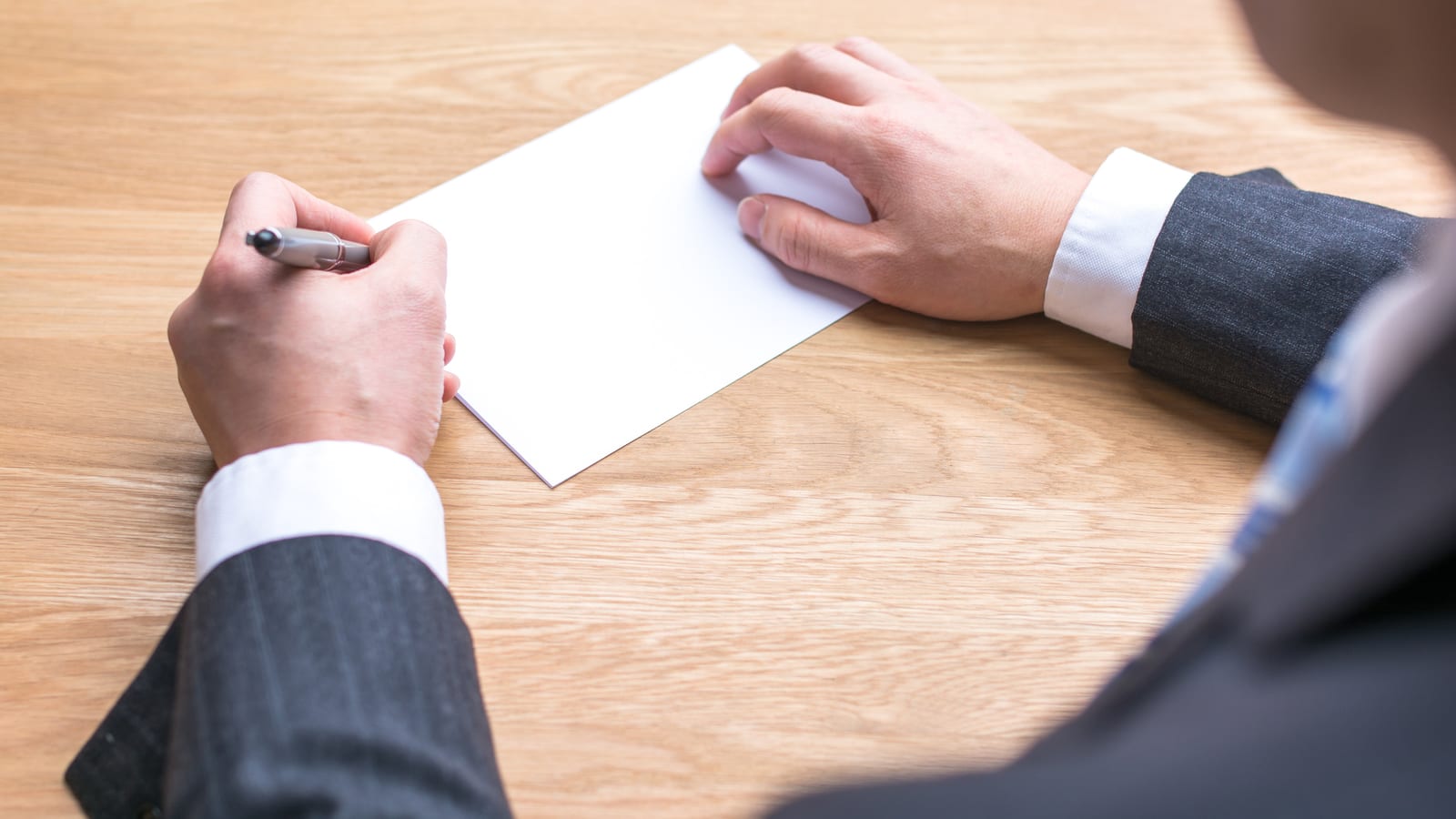 A man in a dark suit and white shirt writing by hand on a plain white card at a wooden desk