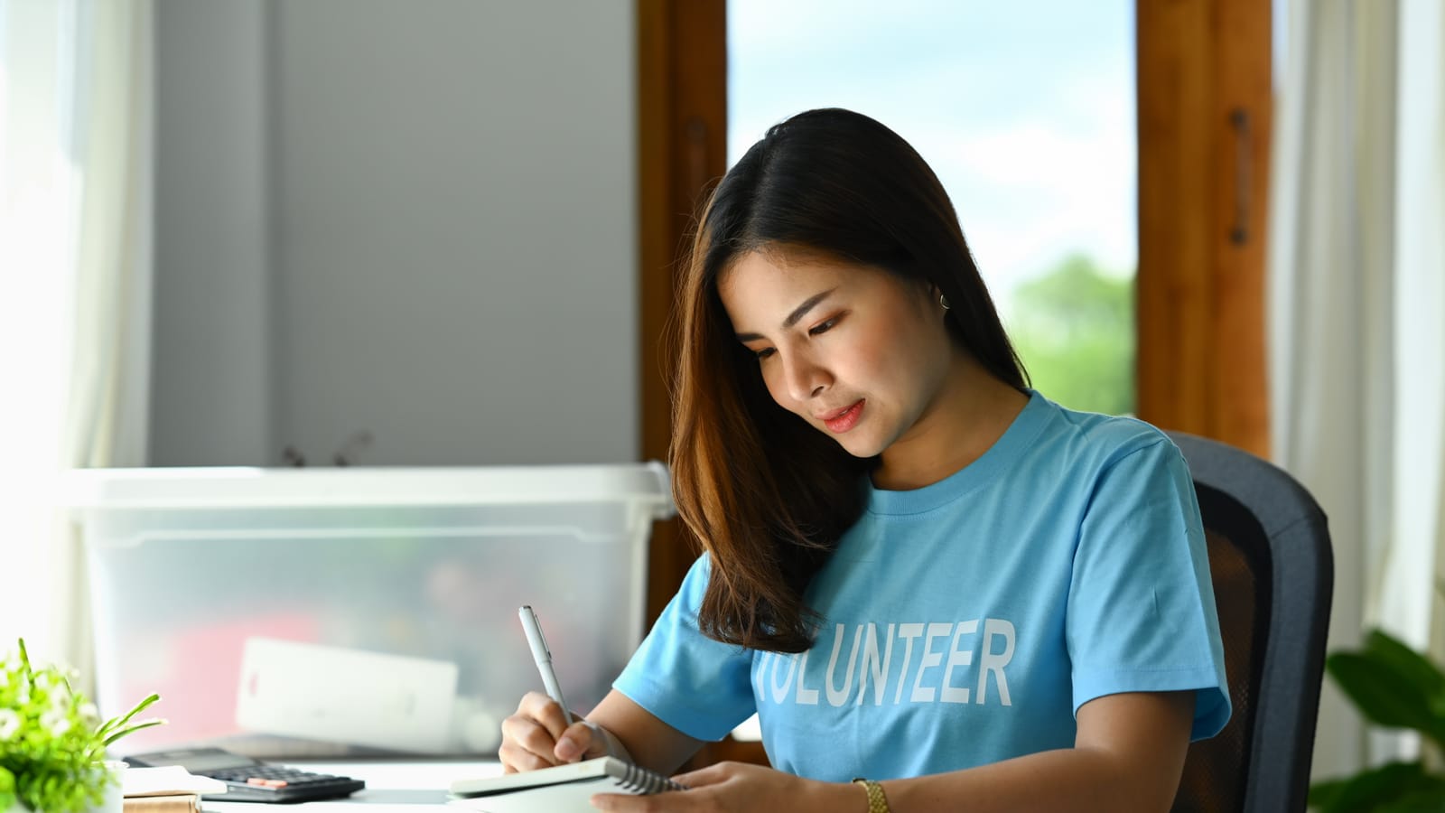 A nonprofit volunteer at a shared office table writing a thank-you card to a donor, a stack of addressed envelopes beside her