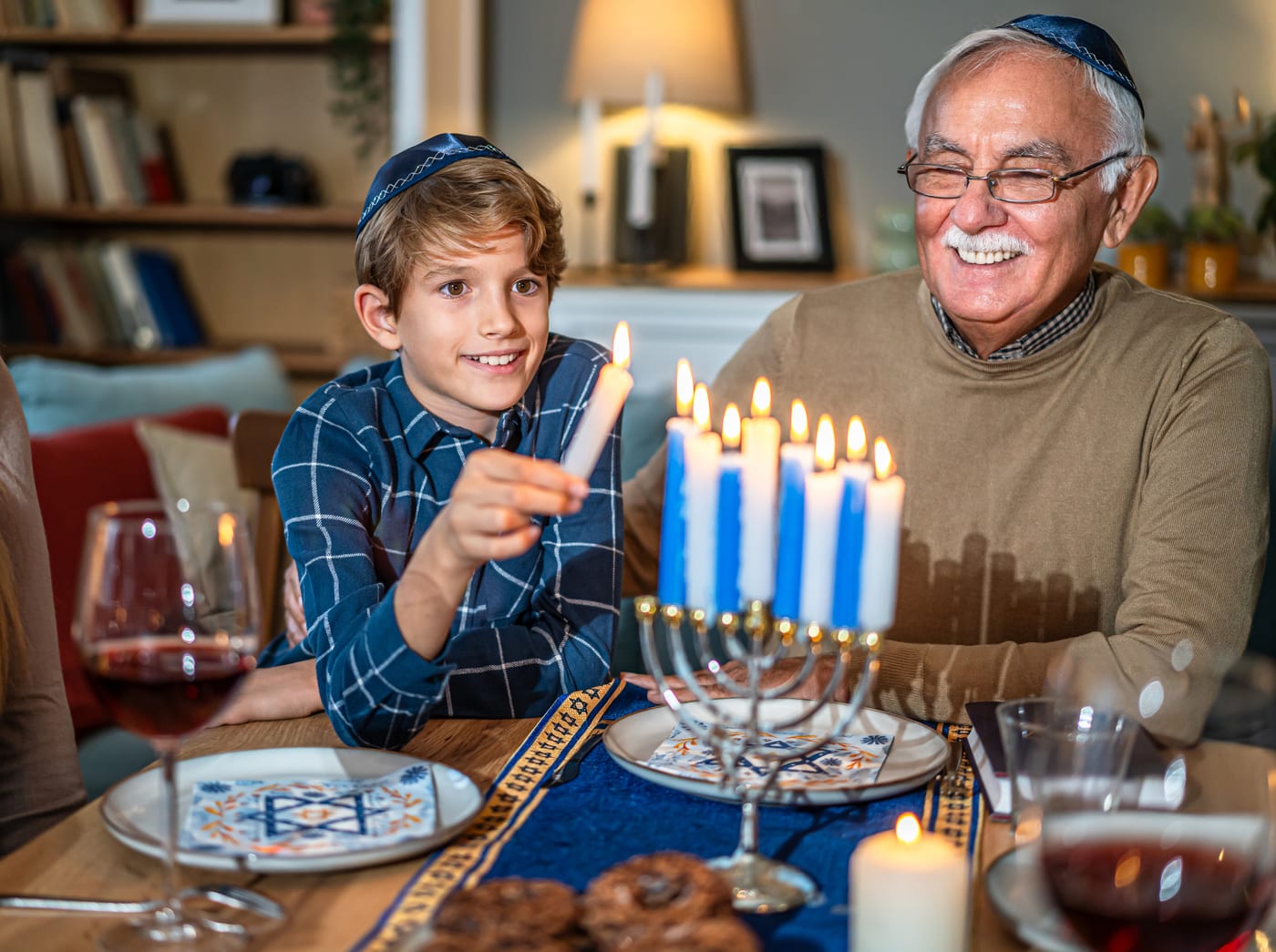 A boy lighting the menorah candles with his smiling grandfather at the holiday table