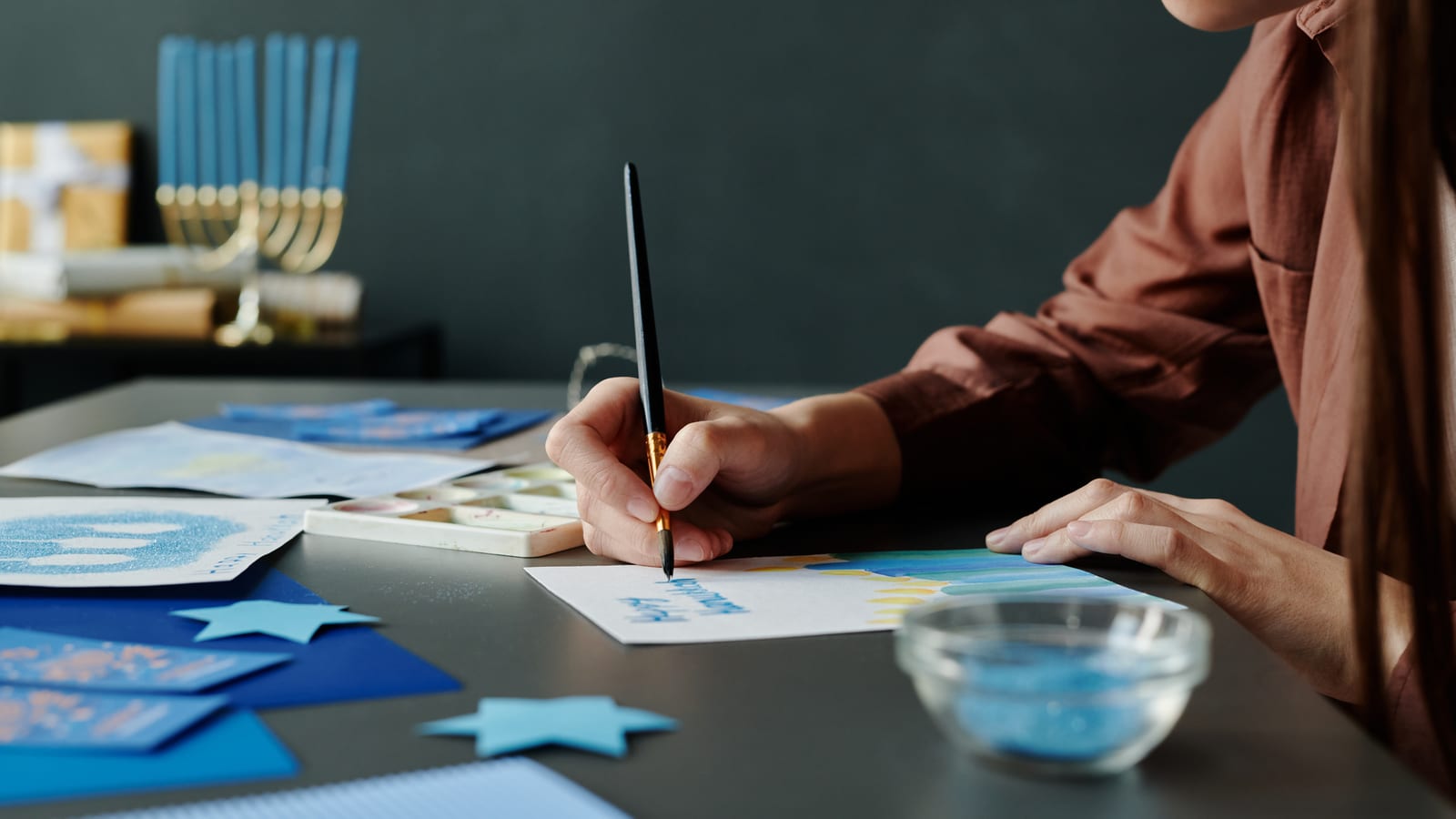 A young woman writing Hanukkah wishes by hand on a handmade card, a menorah blurred behind her