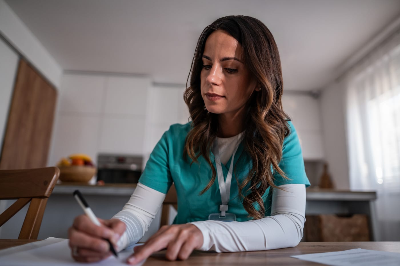 A nurse in teal scrubs sitting at a kitchen table, writing by hand on paper, deep in thought