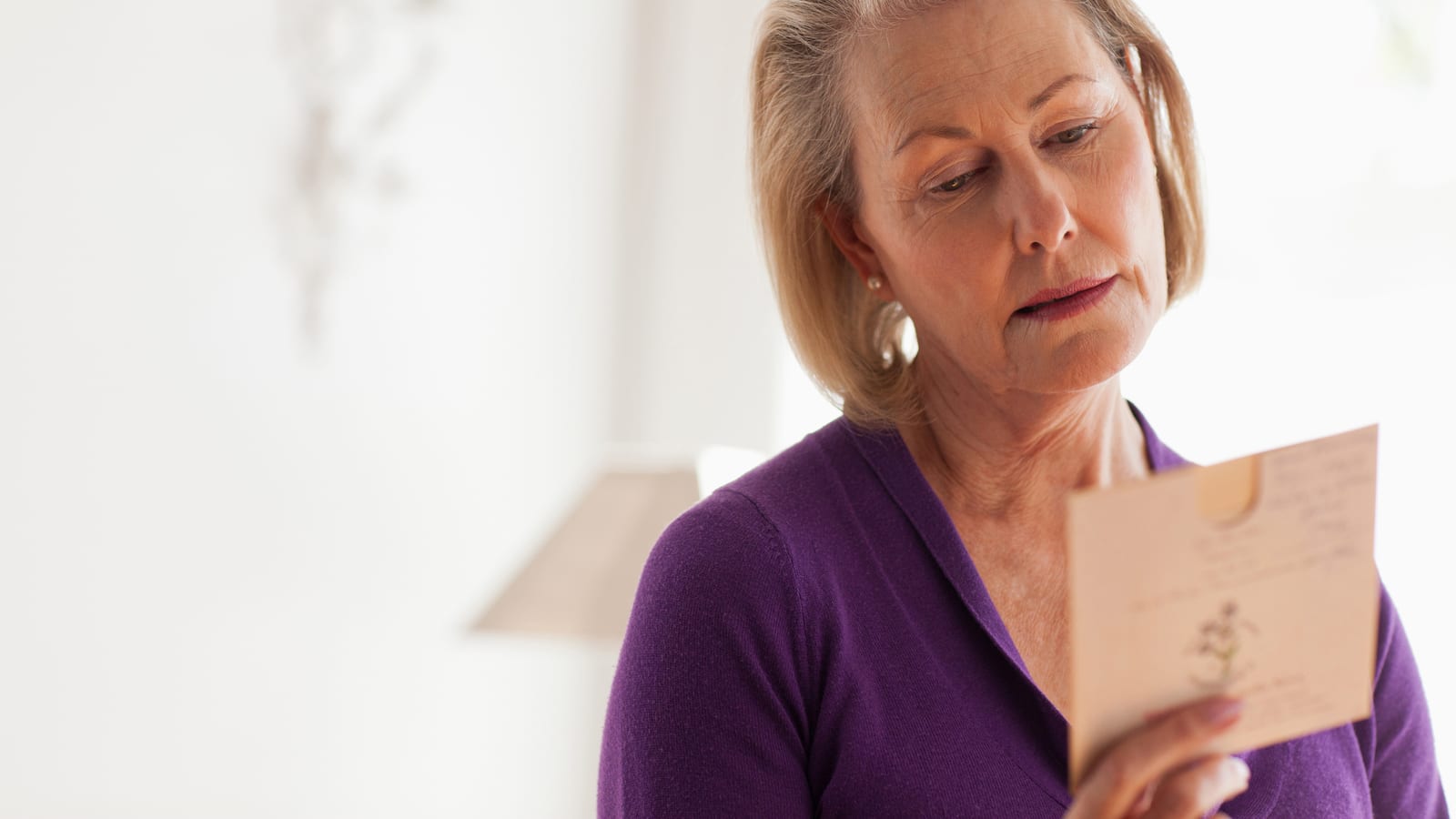 An older woman in a purple top standing quietly at home, reading a card she holds in both hands