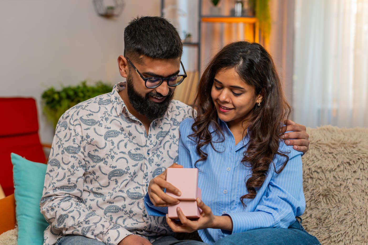 A young Indian couple sitting close on their sofa at home, opening a small pink jewellery box together