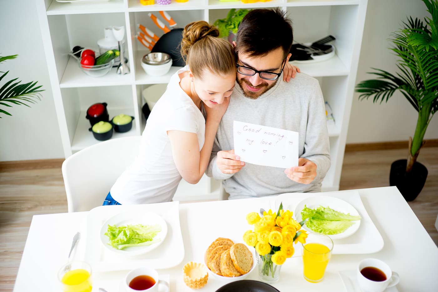 A young couple on the front steps of a house reading a handwritten card together, a set of new keys in one hand