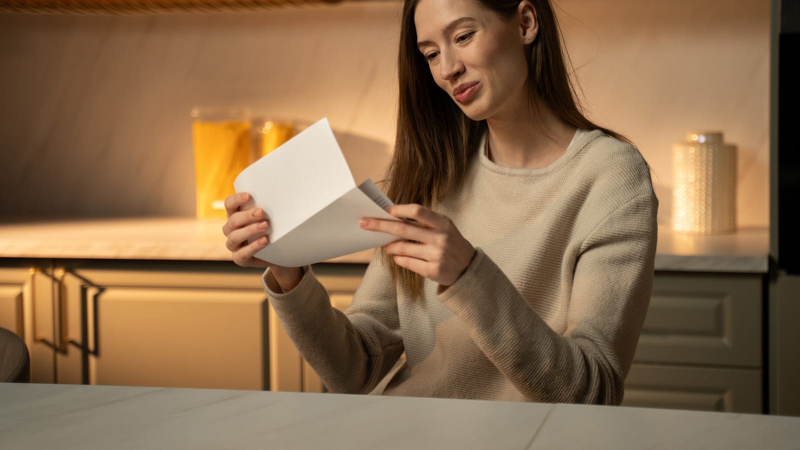 A smiling young woman in a warmly lit kitchen reading a card she has just opened