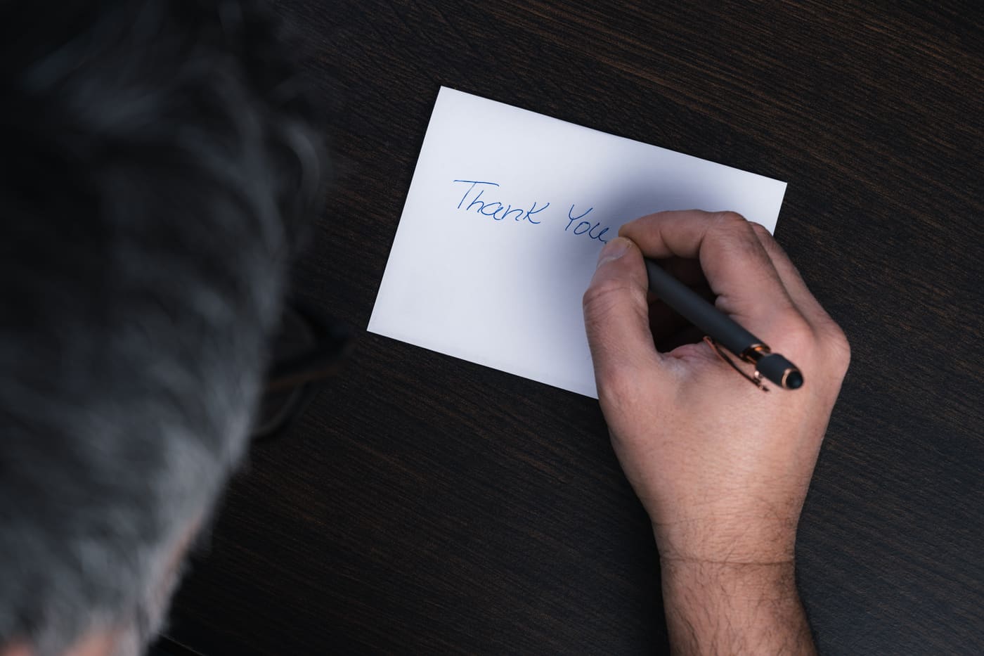 A hand writing Thank You on a plain white card with a black pen, resting on a dark wooden desk