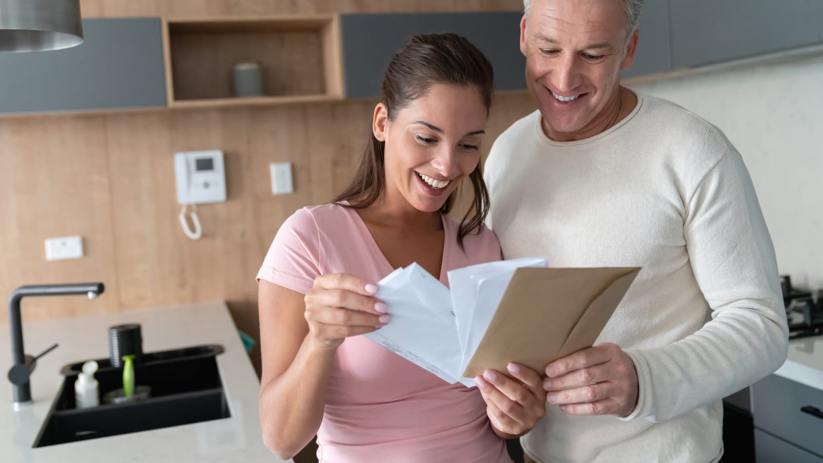 A smiling couple in their kitchen opening a stack of mail together, a brown envelope and a card in their hands