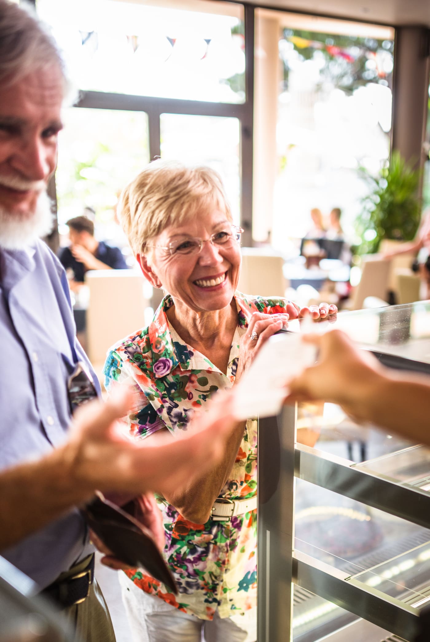 A server handing a handwritten card to a smiling older couple at their usual corner table in a busy neighbourhood restaurant