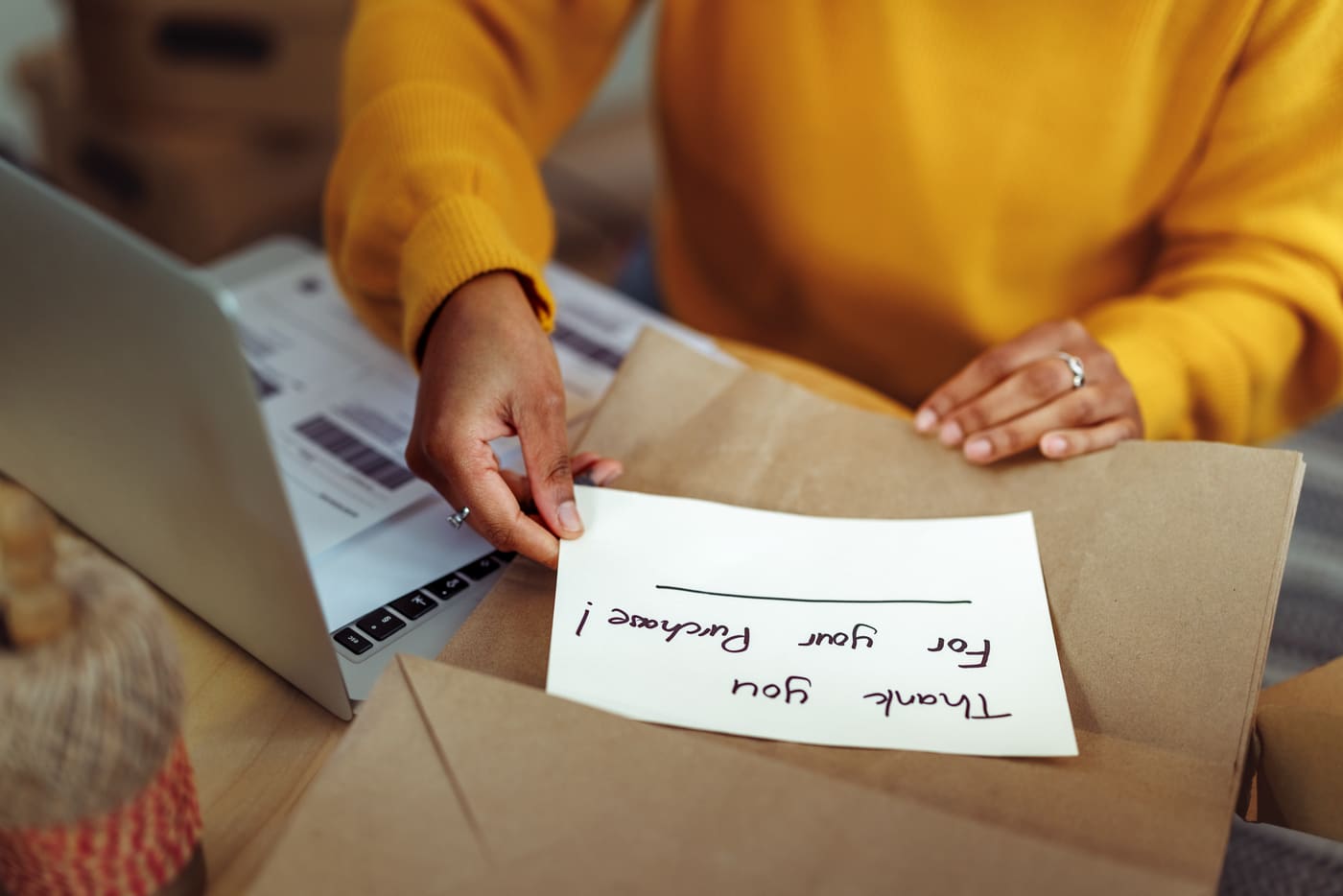 A shop owner tucking a handwritten thank you for your purchase card into a brown paper parcel beside a laptop