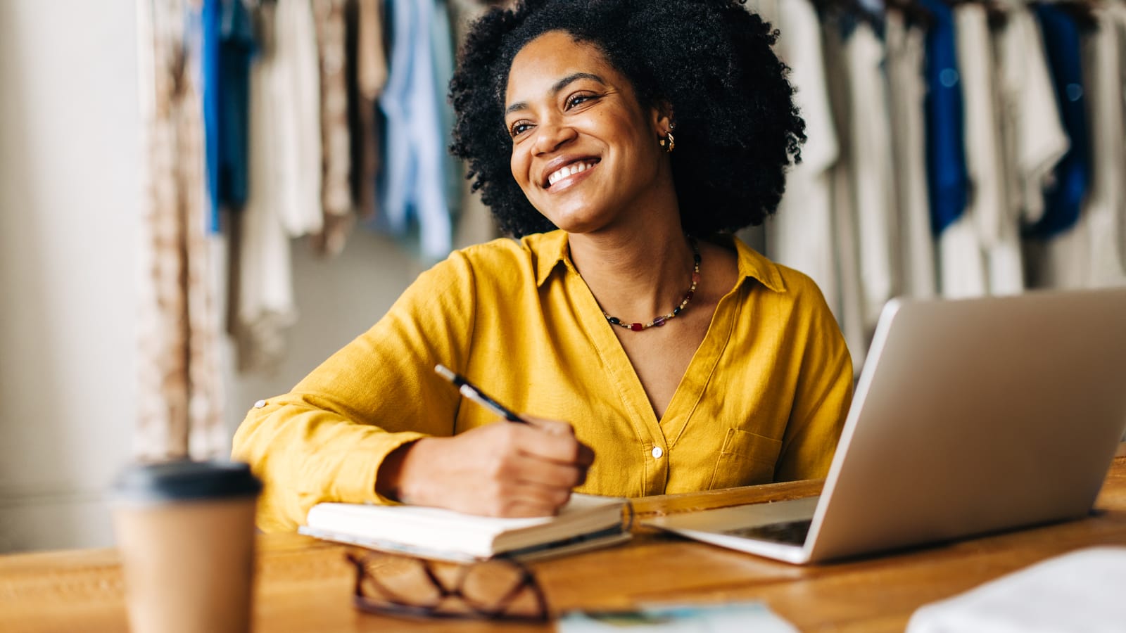 A smiling boutique owner in a yellow shirt writing in a notebook at her table, clothing racks behind her