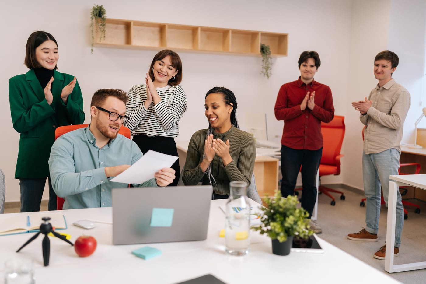 A team clapping round a desk while one colleague, smiling, holds up a sheet of paper