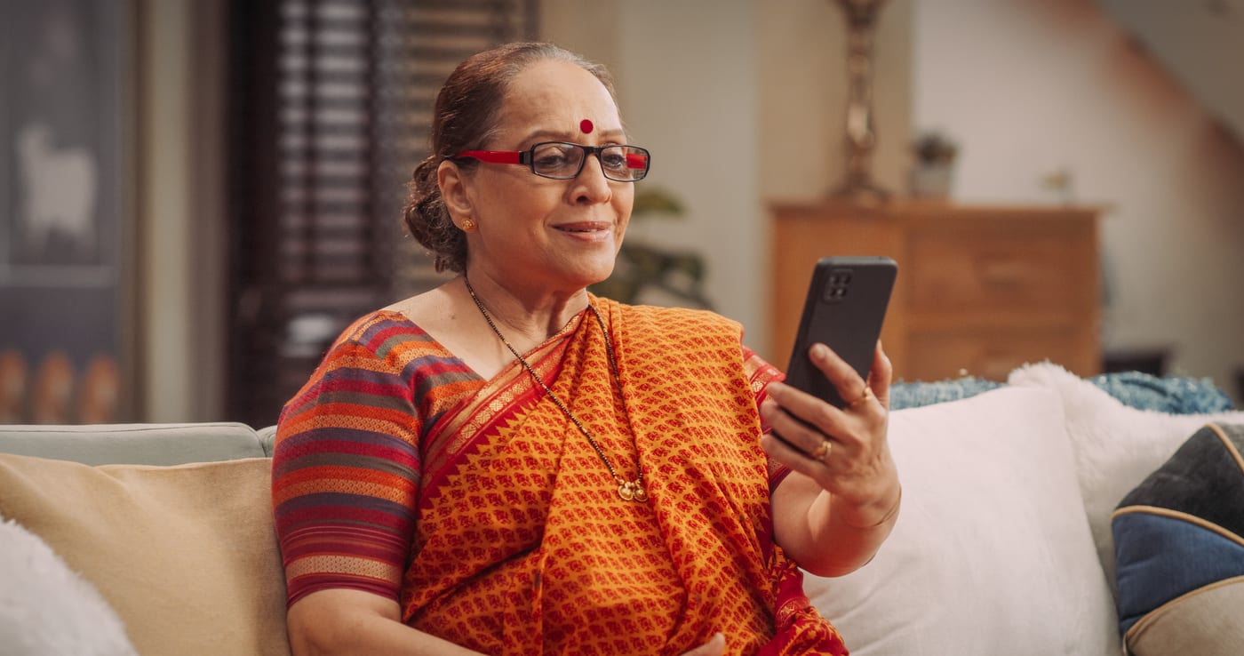 An Indian grandmother in an orange sari and red glasses smiling at her phone on a video call at home