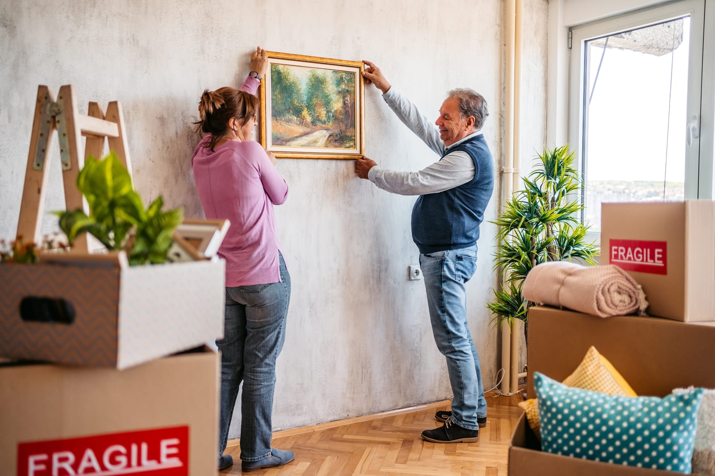 An older couple hanging a framed painting in their new home, moving boxes around them