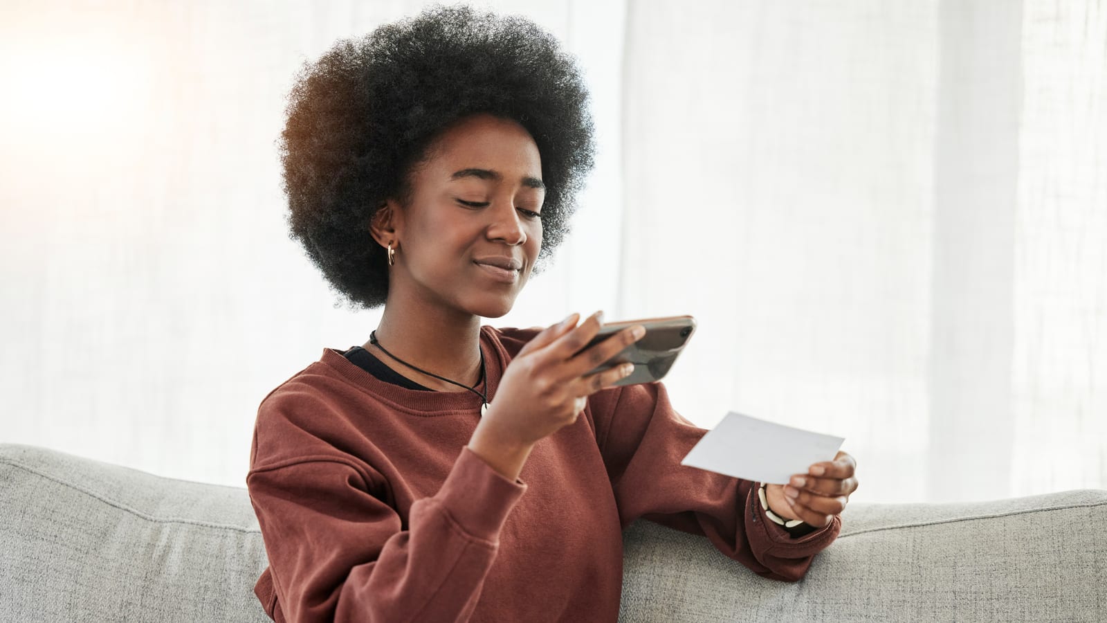 A smiling woman on her sofa holding a card in one hand and photographing it with the phone in her other