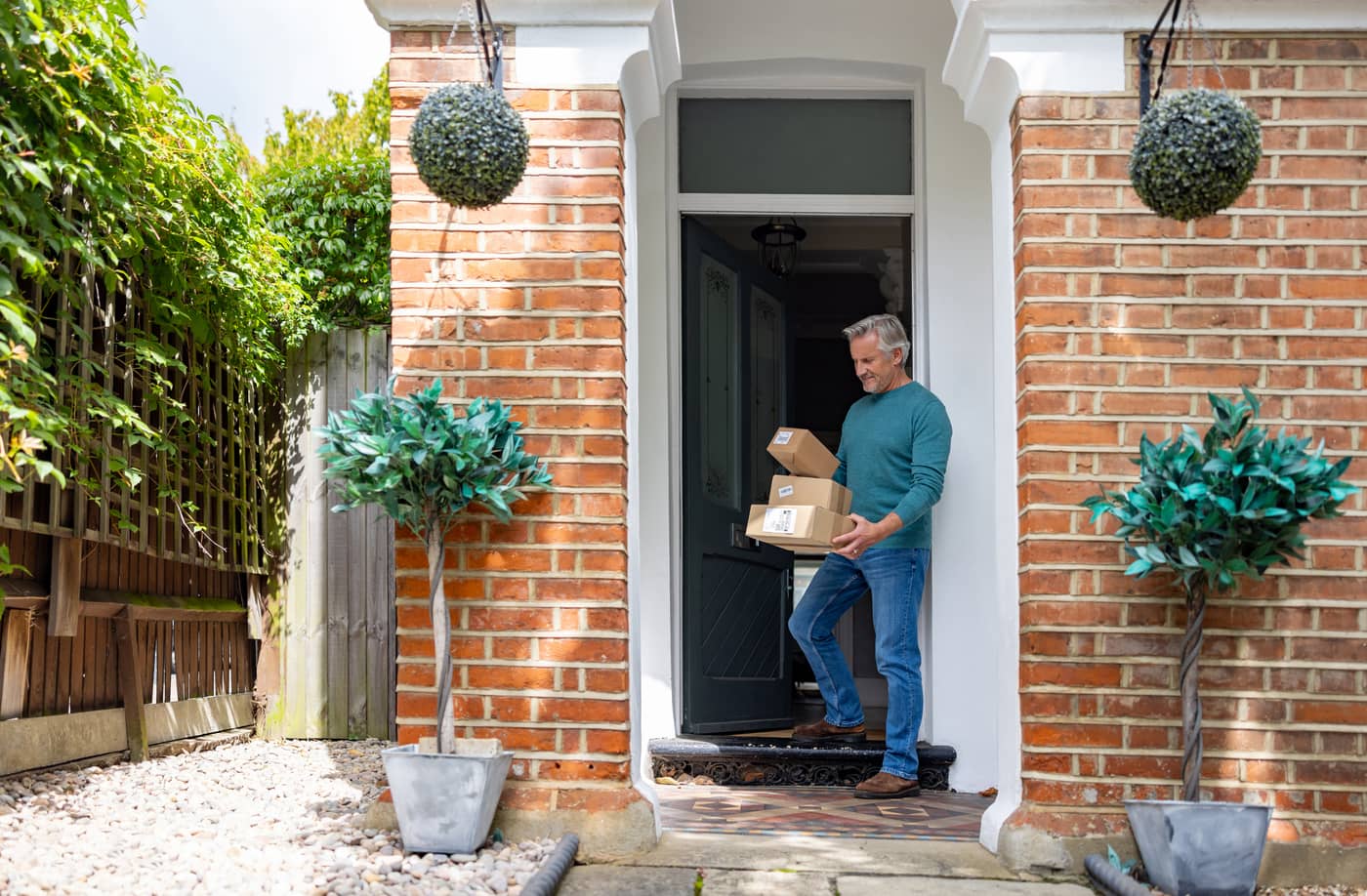 A man stepping out of the front door of his brick house carrying two cardboard parcels in his arms