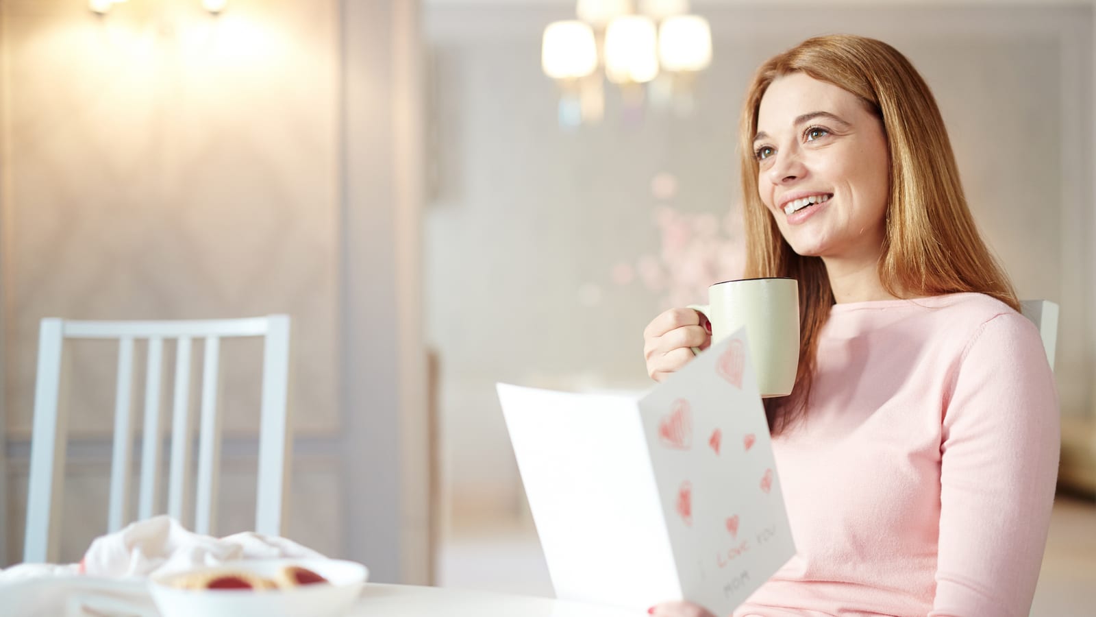 A smiling woman at her table with a mug of coffee in one hand and an open card in the other