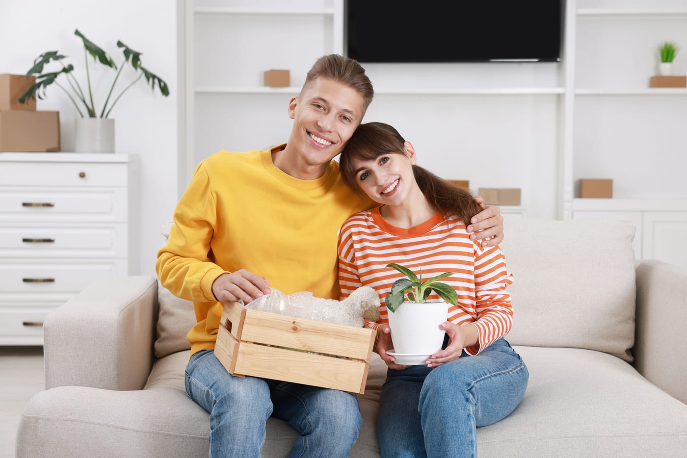 A happy young couple on the sofa of their new apartment holding a crate and a houseplant