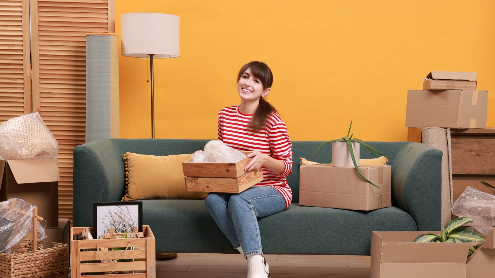 A smiling woman sitting on a green sofa in her new apartment, moving boxes and a wooden crate around her