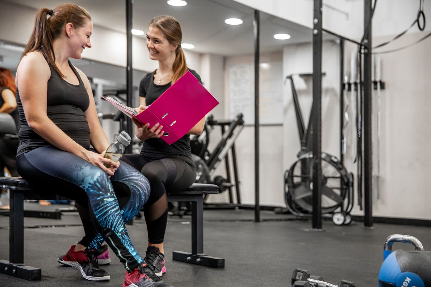 A personal trainer in a bright gym handing a handwritten card to a smiling client beside a squat rack after a session