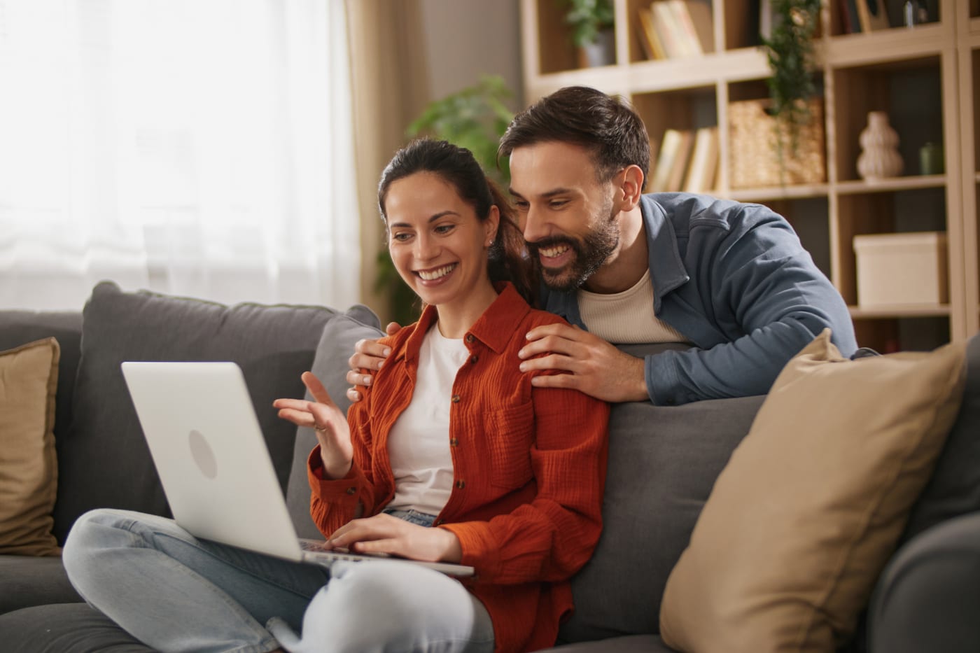 A laughing couple on their grey sofa at home looking at a laptop together, his hands on her shoulders