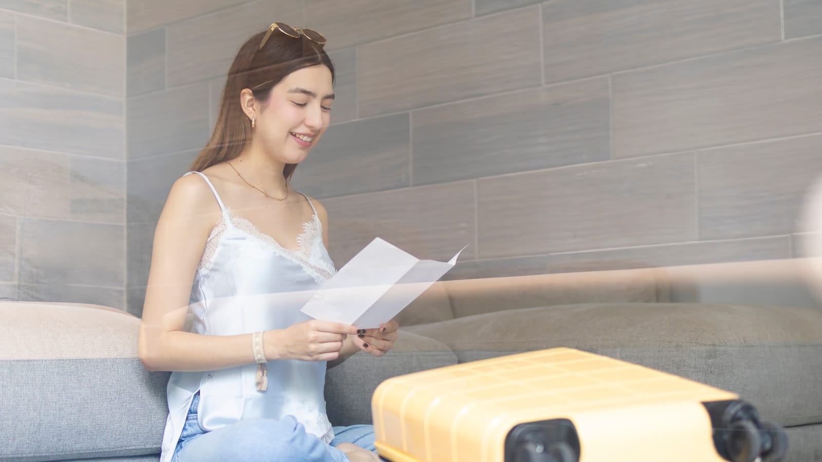 A smiling woman sitting on the floor beside her yellow suitcase, reading a sheet of paper in her hands