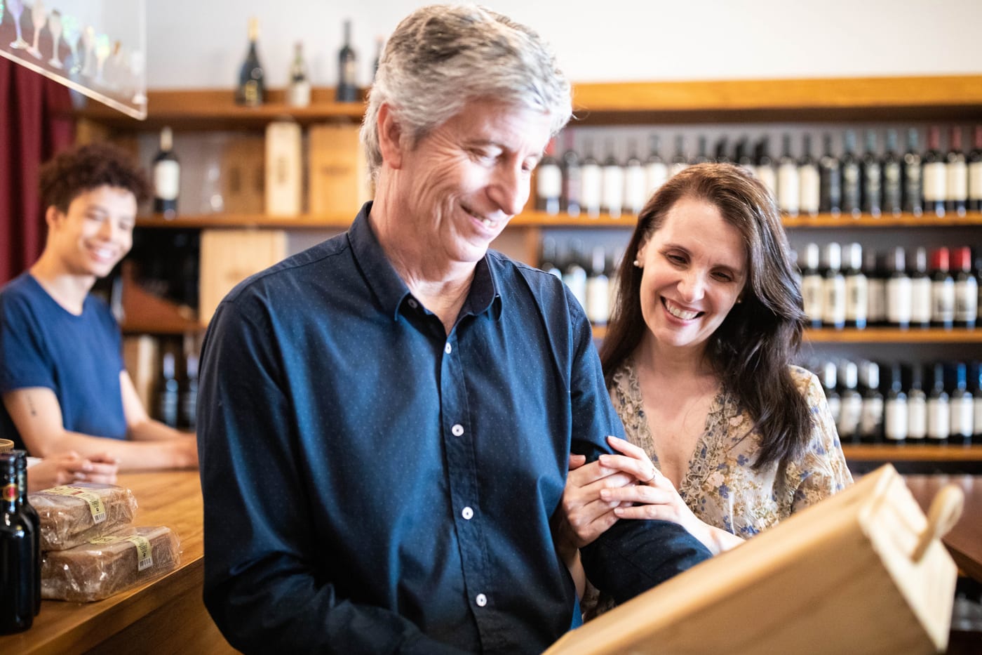 A smiling couple at a wine shop counter looking into a wooden wine box, bottles on the shelves behind them